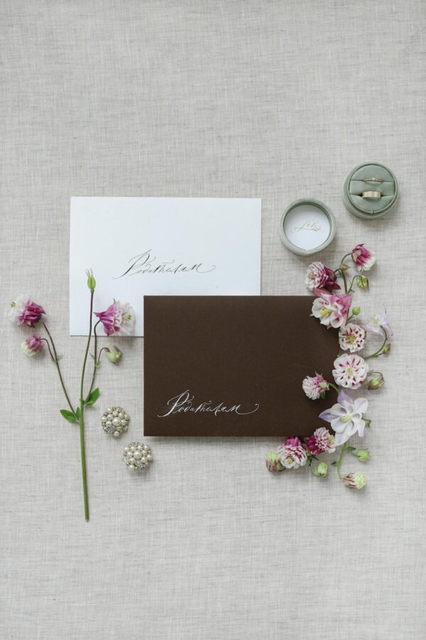 Chic flat lay of wedding invitations, florals, jewelry, and rings on a textured surface.