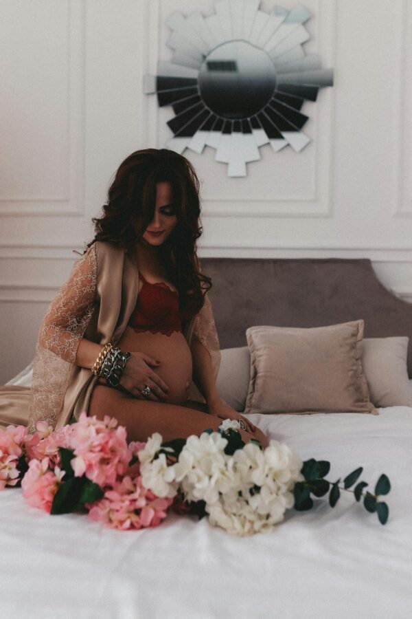 Pregnant woman with long black hair and bracelets sits on bed with floral decor, embracing her belly.
